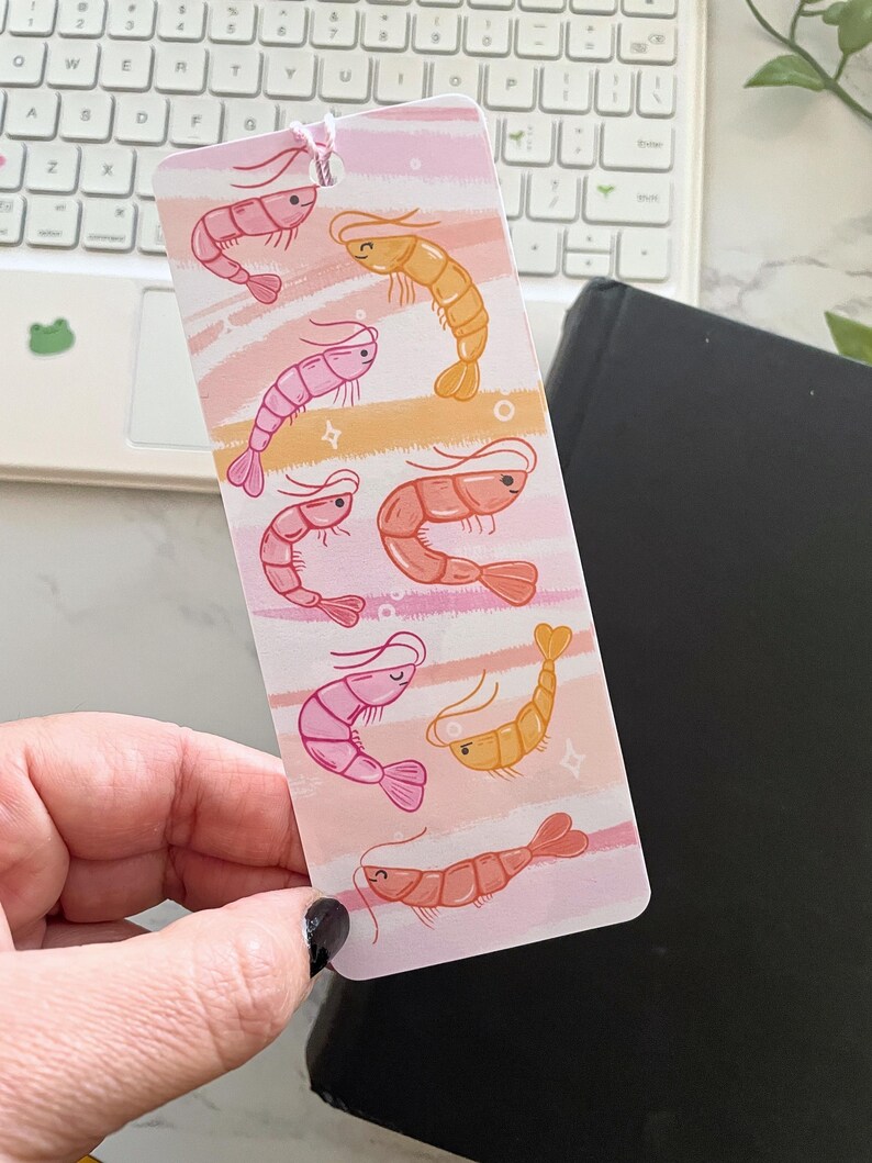 May include: A pink and orange bookmark with a whimsical design of multiple shrimp. The shrimp are illustrated in a cartoon style with different expressions and are set against a background of pink and orange stripes.