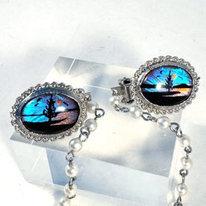 May include: A pair of silver-toned cufflinks with an oval design featuring a landscape scene with a tree, sun, and birds. The cufflinks are connected by a chain of small, white pearls.