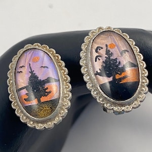 May include: A pair of vintage silver-toned clip-on earrings. Each earring features an oval design with a landscape scene in purple, orange, and black. The earrings have a scalloped edge.
