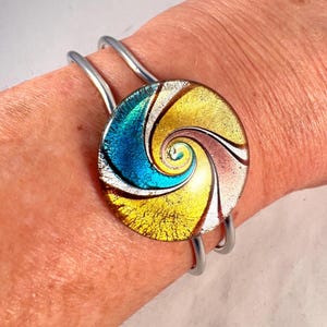 May include: A silver cuff bracelet with a dichroic glass disc. The glass has a spiral pattern in blue, gold, and brown. The bracelet is a fashionable accessory for the wrist.