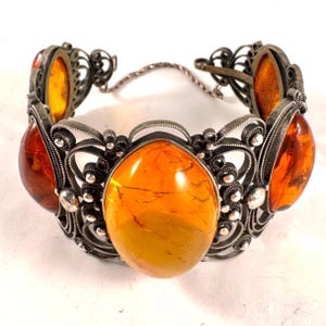Könnte beinhalten: Ein Gliederarmband mit fünf großen, ovalen, durchscheinenden Bernsteinsteinen, die in dunkles, oxidiertes Silberfiligran gefasst sind. Das Armband weist detaillierte Drahtverzierungen und eine dünne Sicherheitskette aus Silber auf.