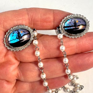 May include: Vintage silver cufflinks with an oval design depicting a landscape scene with a tree, birds, and a sunset. The cufflinks are linked by a delicate chain of small, white pearls.