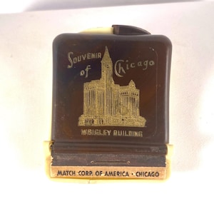 Könnte beinhalten: Ein rechteckiges Streichholzschachtel-Etui aus braunem Kunststoff mit cremefarbener Basis. Die Vorderseite zeigt eine goldfarbene Grafik des Wrigley Building mit dem Text Souvenir of Chicago. Der untere Streifen zeigt MATCH CORP. OF AMERICA - CHICAGO.