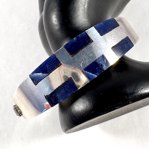 May include: A silver-toned hinged bangle bracelet featuring a geometric pattern of rectangular dark blue stone inlays. The bracelet has a polished metallic finish and a small integrated clasp mechanism visible on the side of the band.