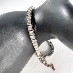 May include: A silver-toned bracelet with square-cut, clear gemstones. The bracelet is composed of a series of connected square links, each holding a single gemstone. A delicate chain extends from the clasp. The bracelet is displayed on a black mannequin arm.