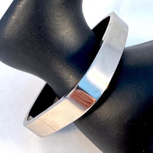 May include: A silver-colored, square-shaped bangle bracelet. The bracelet is shown on a black display stand. The bangle has a smooth, reflective surface, and the design is simple and modern. The bracelet is a piece of jewelry.