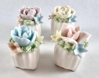May include: Four small, white, fluted ceramic containers, each topped with a cluster of pastel-colored sculpted flowers in shades of light blue, soft pink, pale yellow, and lavender, accented with small green textured foliage.