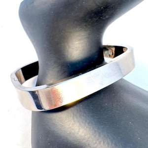 May include: A polished silver-tone metal bangle bracelet with a smooth, flat, rectangular profile. The bracelet features a hinged opening mechanism with a visible clasp on the side, showcasing a clean, reflective surface and a rigid, structured form.