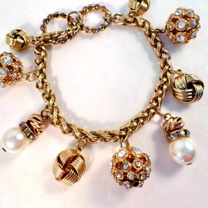May include: Gold-toned charm bracelet featuring a chunky chain design. The bracelet is adorned with various charms, including faux pearls, crystal-studded spheres, and woven knot-shaped charms. The bracelet has a lobster clasp closure.