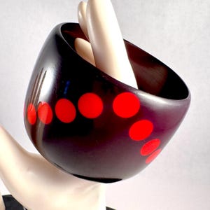 May include: A wide, dark brown, polished bangle bracelet featuring a curved, diagonal row of vibrant, solid red circular dots. The bracelet has a smooth, glossy finish and a thick, rounded profile that encircles the wrist area.
