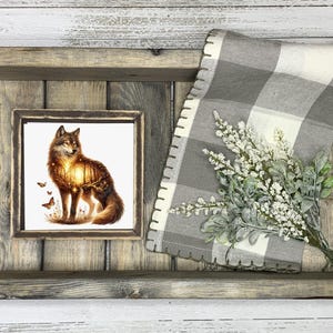 May include: A wooden tray with a framed print of a wolf standing in a forest. The wolf is silhouetted against a sunset. The tray is decorated with a gray and white checkered fabric and a sprig of white flowers.