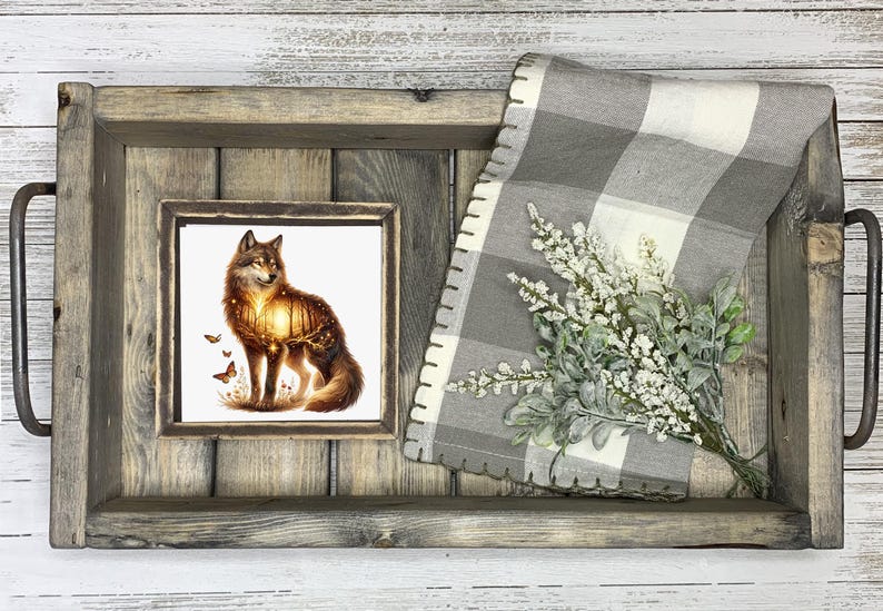 May include: A wooden tray with a framed print of a wolf standing in a forest. The wolf is silhouetted against a sunset. The tray is decorated with a gray and white checkered fabric and a sprig of white flowers.