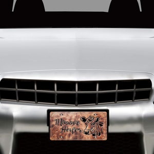 May include: A brown leather-like license plate with a cow head silhouette and the text "Mooove Heifer". The license plate is attached to the front of a silver car.