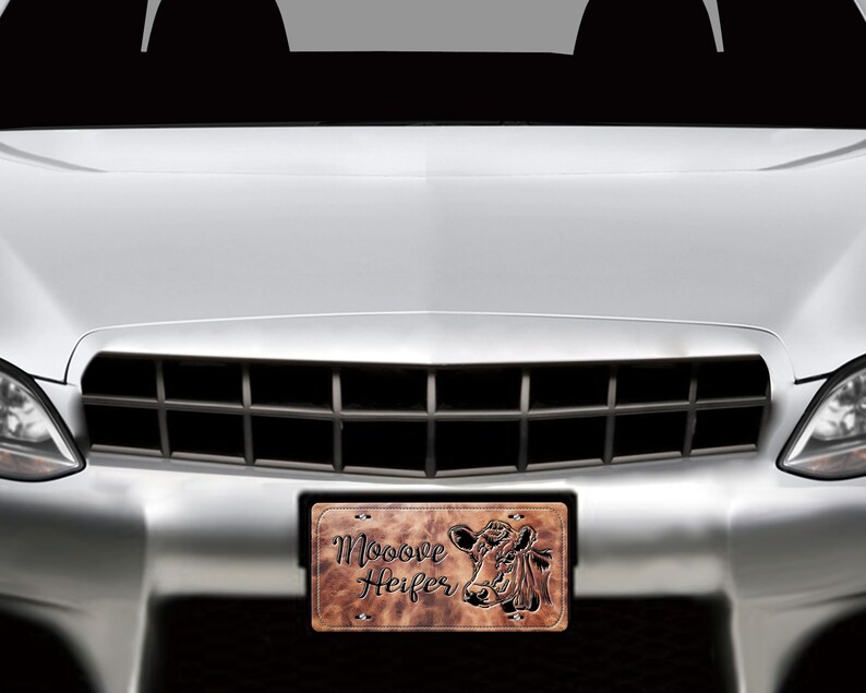 May include: A brown leather-like license plate with a cow head silhouette and the text "Mooove Heifer". The license plate is attached to the front of a silver car.