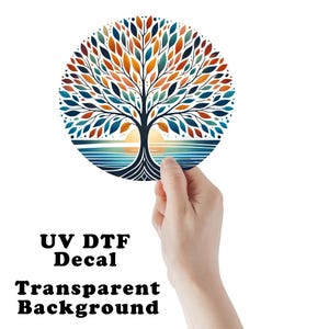 May include: A round UV DTF decal with a transparent background. The design features a stylized tree with colorful leaves in shades of orange, blue, and green. The tree is set against a backdrop of a sunset over water.