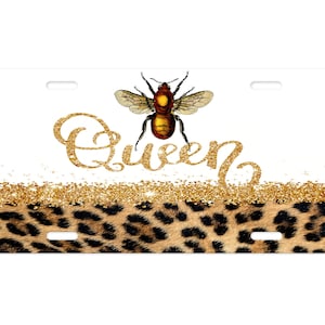 May include: A white license plate with a gold glitter background and a leopard print design at the bottom. The text "Queen" is written in gold glitter with a bee illustration above the text.