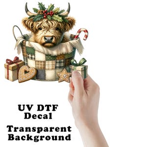 May include: A UV DTF decal featuring a fluffy Highland cow with holly on its head, sitting in a patchwork basket with a candy cane, gift boxes, and gingerbread cookies. The design includes the text UV DTF Decal Transparent Background.