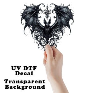 May include: Black bat decal with ornate wing and body details, held against a white background. The decal has the text "UV DTF Decal" and "Transparent Background". The bat design features intricate patterns and a gothic aesthetic.