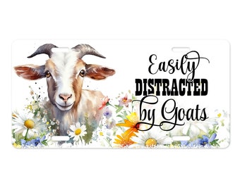 Puede incluir: Una ilustración de acuarela de una cabra con pelaje blanco y marrón rodeada de flores coloridas. El texto "Easily Distracted by Goats" está escrito en cursiva negra.