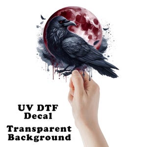 May include: A black raven with red eyes is perched in front of a large, red moon. The raven is detailed with textured feathers. The words "UV DTF Decal" and "Transparent Background" are below the raven.