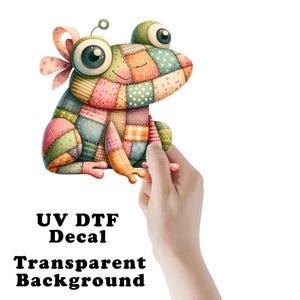 May include: A colorful frog decal with a patchwork design. The frog features large, expressive eyes and a whimsical bow. The text "UV DTF Decal Transparent Background" is printed below the frog.