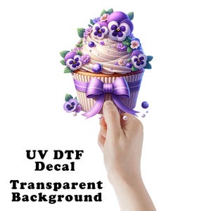 May include: A cupcake with purple and white frosting, decorated with purple pansies and blueberries. The cupcake is on a white background with a purple ribbon and a hand holding it. The text "UV DTF Decal Transparent Background" is below the cupcake.
