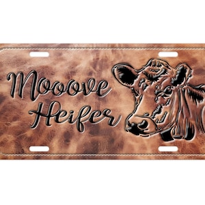 May include: A brown leather license plate with a black stitched border. The plate features a black silhouette of a cow's head and the text "Mooove Heifer".