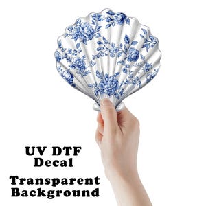 May include: A white and blue floral seashell-shaped decal. The decal features a detailed blue floral pattern on a white background. The text "UV DTF Decal Transparent Background" is printed below the seashell.