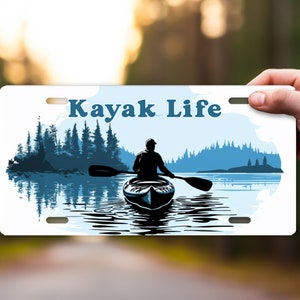 May include: A white license plate with the words "Kayak Life" in blue. The plate features a silhouette of a kayaker on a lake with trees in the background. The design is in shades of blue and white.
