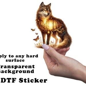 May include: A golden wolf with a forest scene inside its body, surrounded by butterflies. The wolf is being held by a hand. The text "Apply to any hard surface Transparent Background UV DTF Sticker" is below the image.