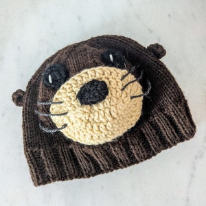 Pode incluir: Um chapéu de malha marrom com um rosto de lontra bege e preto na frente. O chapéu tem uma borda de malha marrom e um topo de malha marrom.