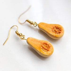 May include: A pair of gold dangle earrings with a halved butternut squash charm. The squash is painted a light orange colour with brown seeds.