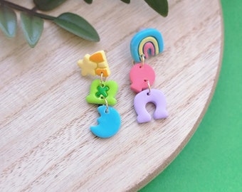 May include: Colorful polymer clay earrings featuring a rainbow, horseshoe, moon, and clover design. The earrings have a playful, whimsical style with bright colors including blue, pink, green, yellow, and purple. The earrings are displayed on a wooden surface.