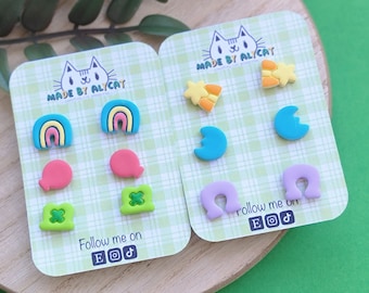 May include: Two sets of colorful stud earrings on cards. One set features rainbow, clover, and balloon shapes. The other set includes star, crescent moon, and horseshoe shapes. The cards have a cat logo and the text "MADE BY AL/CAT" and "Follow me on".