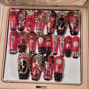 Könnte beinhalten: Ein Set roter Kunstnägel mit verschiedenen Designs, darunter Champagnergläser, Uhren und dem Text "Happy New Year". Die Nägel sind mit Strasssteinen und schwarzen Perlen verziert, was einen festlichen und glamourösen Look erzeugt.