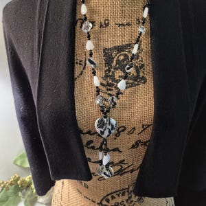 May include: A black and white beaded necklace featuring various stone shapes, including heart-shaped pendants. The necklace is displayed on a mannequin wearing a black bolero jacket.