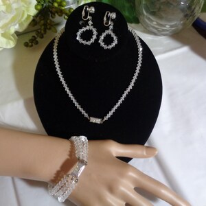 May include: A set of crystal jewellery featuring a necklace, bracelet, and earrings. The necklace and bracelet are made of small, clear crystals strung together. The earrings are teardrop-shaped and have a clip-on fastening.
