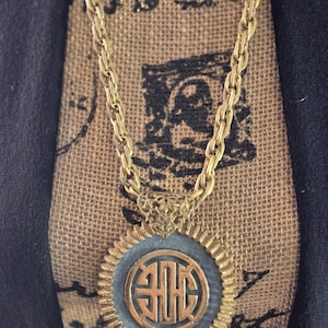May include: A gold-coloured necklace with a circular pendant. The pendant has a central design within a textured frame. The chain is a thick, linked style. The necklace is displayed against a textured, neutral background.
