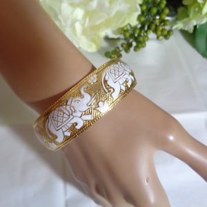 May include: Gold-tone bangle bracelet with white elephant designs. The bracelet features a repeating pattern of elephants and floral accents. The bracelet is approximately 2 inches wide.