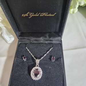 May include: A jewelry set featuring a silver-toned necklace with an oval-shaped dark red gemstone pendant surrounded by small clear stones, and matching stud earrings. The set is presented in a black box with the text 14k Gold Plated inside.
