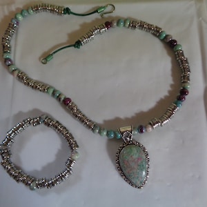 May include: A silver necklace with a green and pink stone pendant and a matching silver bracelet. The necklace has a green cord with a silver clasp.
