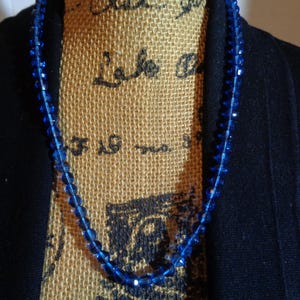 May include: A blue glass bead necklace with a simple design. The necklace is on a hessian mannequin.