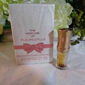 May include: A small, clear glass bottle with a gold cap filled with a light brown liquid. The bottle is labeled "The Perfume of Fleurs D'Elle, One-Eighth Ounce Spray Mist, Nettie Rosenstein, Inc., Dist. New York, N.Y. 10019".