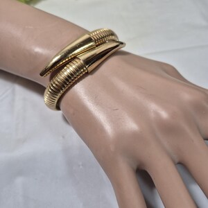 May include: A gold-toned cuff bracelet with a textured, ribbed design. The bracelet features two pointed, horn-like ends that overlap. The jewelry is displayed on a mannequin hand, showcasing its design and style.