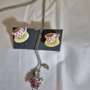 May include: A silver necklace with a teapot pendant and a pair of earrings. The earrings and the pendant are ceramic teacups with purple polka dots and a yellow saucer. The teapot pendant is pink.