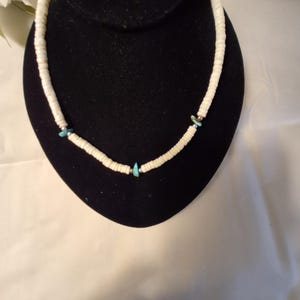 May include: A white shell necklace with turquoise accents. The necklace is displayed on a black velvet bust. The necklace features small, flat, white shell beads strung together, with turquoise-colored stones interspersed throughout the design. The necklace is a choker style.