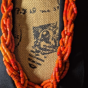 May include: A multi-strand necklace composed of small, cylindrical orange beads arranged in a braided pattern. The necklace is displayed against a textured tan fabric background featuring black printed text and a partial black graphic design.