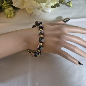 May include: A bracelet featuring round black beads with a floral pattern in shades of pink, white, and green, interspersed with small gold-toned square spacers. The bracelet is secured with a gold-toned toggle clasp closure.