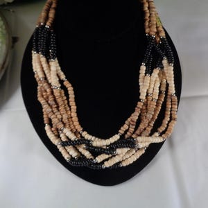 May include: Multi-strand necklace made of wooden beads in shades of brown and black. The necklace is layered and features a variety of bead sizes and shapes.