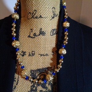 May include: A gold and blue beaded necklace with gold accents. The necklace is on a hessian mannequin.